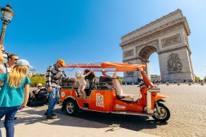 Orange tuk tuk with passengers beside the Arc de Triomphe in Paris