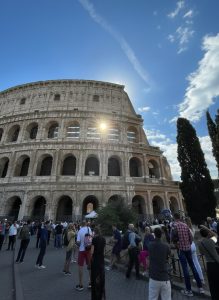 Visitors gathered outside the Colosseum in Rome beneath a bright blue sky with the afternoon sun shining through the ancient arches.