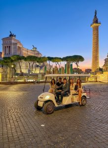 Guests enjoying an evening golf cart tour through Rome with Trajan's Column and Piazza Venezia illuminated in the background.
