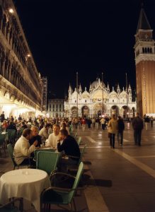 Evening view of St. Mark's Square in Venice with outdoor cafés, visitors, St. Mark's Basilica, and the Campanile illuminated at night.